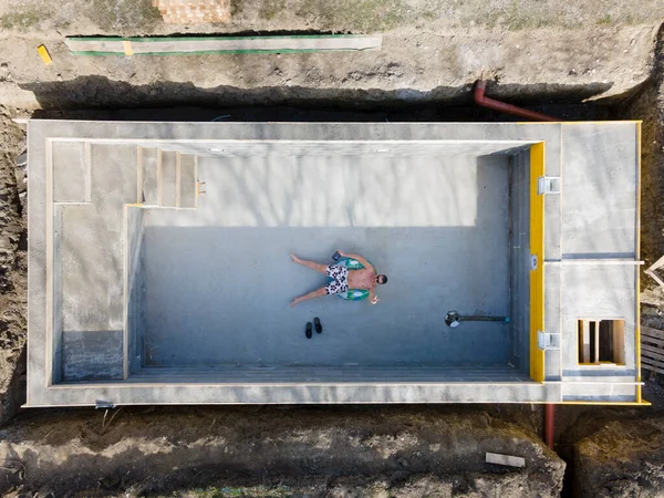 穿着泳裤的男人躺在一个漂浮的轮胎上的尚未完工的游泳池里 用无人驾驶飞机自杀 — 图库照片