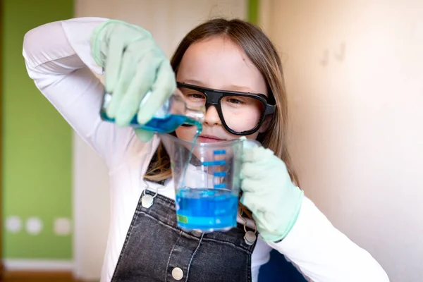 pretty young schoolgirl sitting on her desk in room at home experimenting with blue liquid while homeschooling chemistry during corona time