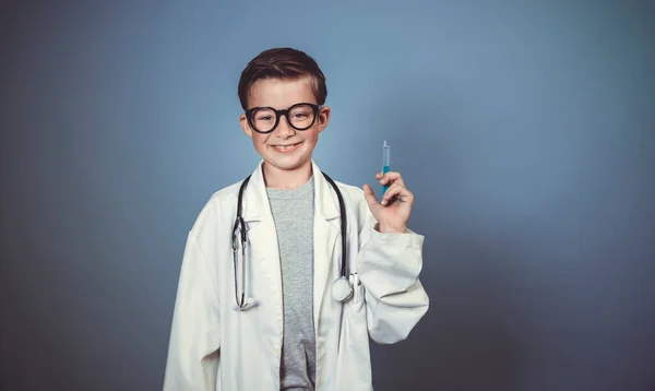 クールな少年は白い医者のスモックを持つ医者として偽装されており 青い背景の前で注射器と聴診器で遊ぶ — ストック写真