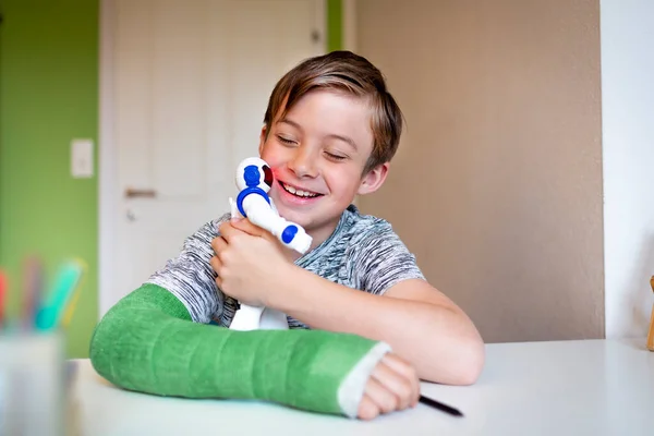 cool boy with green arm cast is sitting in his room and is playing with toy robot
