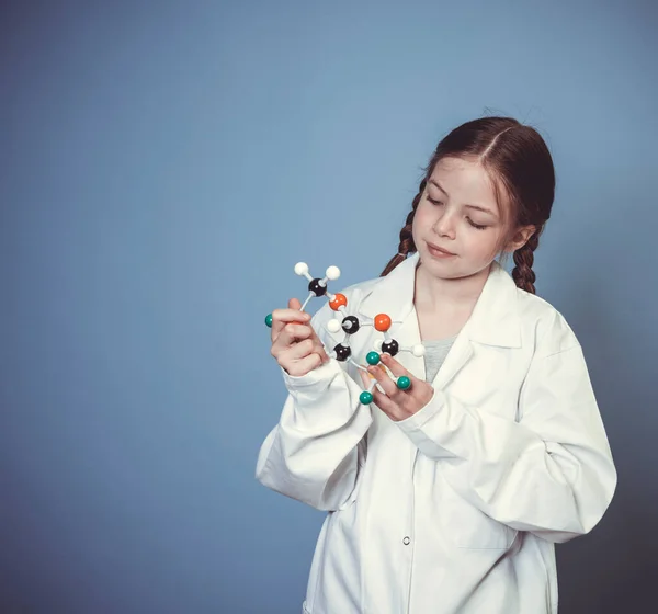 2本のブレイドを持つ可愛い女の子は科学者として服を着て青い背景の前で分子のモデルを手にしています — ストック写真