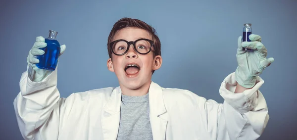濃い黒メガネのクールな少年は白いコートを着た科学者として服を着て青い背景の前で青い液体を実験しています — ストック写真