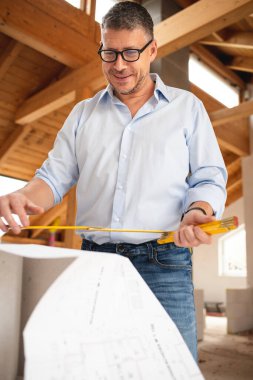 Siyah gözlüklü ve mavi gömlekli adam plana bakıyor ve çatı katındaki inşaat çalışmalarını kontrol ediyor.