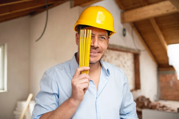 Architect with yellow safety helmet, blue shirt and jeans checks construction progress on building site in loft, attic with a yellow folding rule