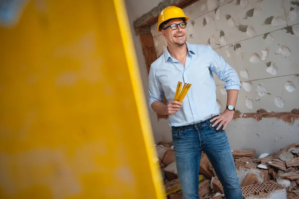 Architect with yellow safety helmet, blue shirt and jeans checks construction progress on building site in loft, attic with a yellow folding rule