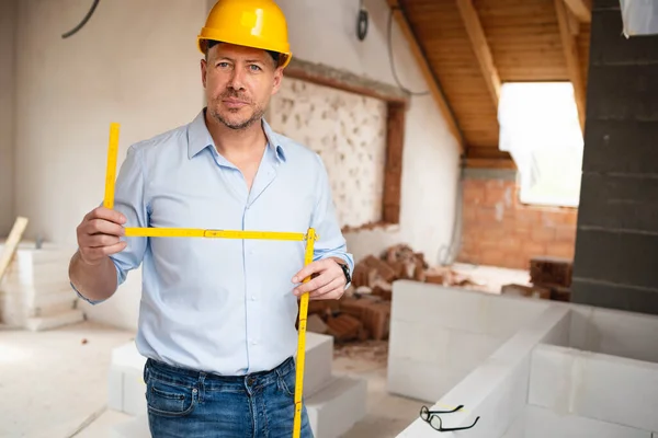 Architect with yellow safety helmet, blue shirt and jeans checks construction progress on building site in loft, attic with a yellow folding rule