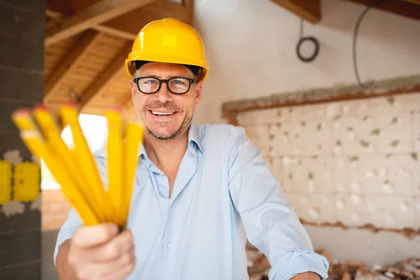 Architect with yellow safety helmet, blue shirt and jeans checks construction progress on building site in loft, attic with a yellow folding rule