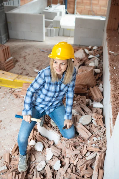 pretty young female worker with blue work shirt and yellow protective helmet works on construction site and holds hammer in her hand, concept of female worker