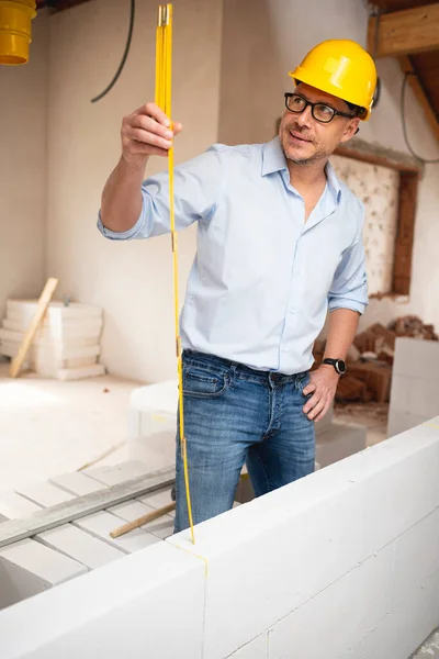 Architect with yellow safety helmet, blue shirt and jeans checks construction progress on building site in loft, attic with a yellow folding rule