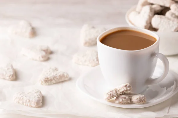 Photo of a vegan breakfast. White cup of coffee with almond milk. Vegan coconut cookies are nearby. Heart-shaped white cookies. On a white background close up.