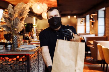 Coronavirüs salgını sırasında dışarıdan yemek. Koruyucu maskeli garson, tıbbi maske takan müşteriye yemek paketi veriyor. Koronavirüs salgını sırasında asgari temas. Sosyal uzaklık.