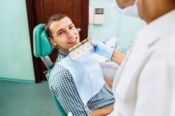 Красивый европейский мужчина улыбается здоровыми белыми и чистыми зубами. Набор имплантатов в разных тонах. Сравнение оттенков зуба с примером. Концепция стоматологической помощи.