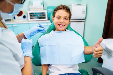 Küçük sabırlı çocuk dişçiye gittiği için mutlu. Acısız diş tedavisi kavramı. Güzel, geniş bir gülümseme bir doktorun muayenehanesinde dişçi koltuğunda oturan bir çocuk. Başparmaklar yukarı..