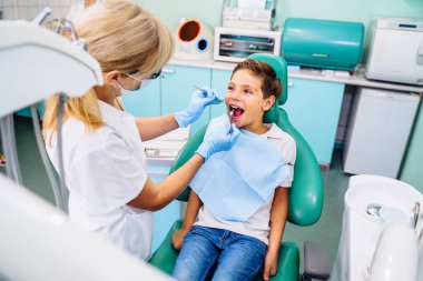 Modern bir klinikte dişçi randevusunda ağzı açık olan küçük bir hasta. Hastanede gülümseyen sevimli çocuk için muayene prosedürü yapan tanınmamış çocuk dişçisinin elleri..