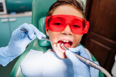 Lambanın ışığıyla ışıklandırma, diş macunuyla. Sınavdaki dişçi muayenehanesindeki genç çocuk. Diş hekimi, klinikte parlak ışık altında diş sandalyesindeki hastaların dişlerini kontrol ediyor..