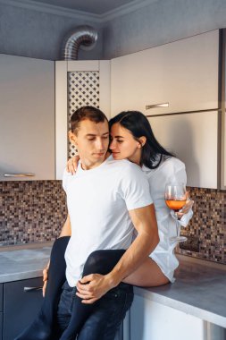 Çekici, seksi, esmer kadın mutfak mobilyalarının üzerinde oturuyor. Bacaklarına tutkuyla sarılan erkek arkadaş. Kadın vücudunu erkeğe bastırıyor. Beyaz gömlekler ve koyu kotlar giymişti..