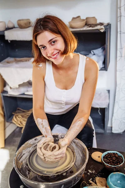 A beautiful young, smiling woman sits on a bench with a potter\'s wheel and makes an earthen pot, vase or mug. Skillfully and accurately draws the correct shape of the product with his hands.