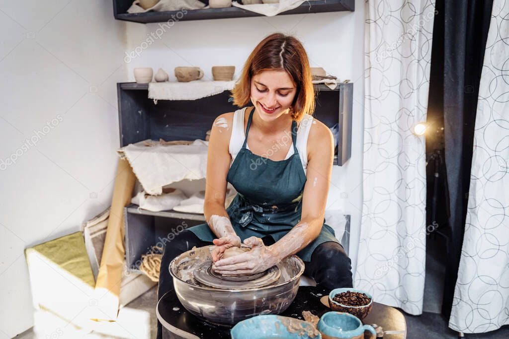 Pottery. A young woman making vase, mug, plate, pot in her own working place. Sitting at the table near the potter's wheel, creating the shape of the future product with his hands.