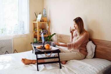 Uzaktan öğrenme, çalışma, online eğitim. Mutlu, güzel bir kadın ofis yerine uzaktan evde yatakta çalışıyor. Bilgisayar, dizüstü bilgisayar ve kahve içerek. Gülümsüyor ve düşünüyor.