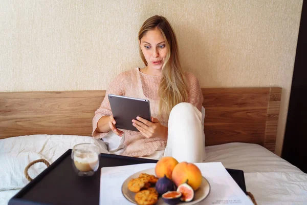 Beautiful young caucasian woman sitting on bed with sleepy look in morning. Working on bed remotely, working from home, busy lifestyle concept. Surfing internet, dressed in casual domestic clothes.