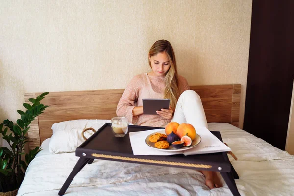 In the morning beautiful caucasian lady sitting and drinking hot tea or coffee in bed. Blond female solving work issues remotely. Cozy workplace at home. Distance learning online education and work.