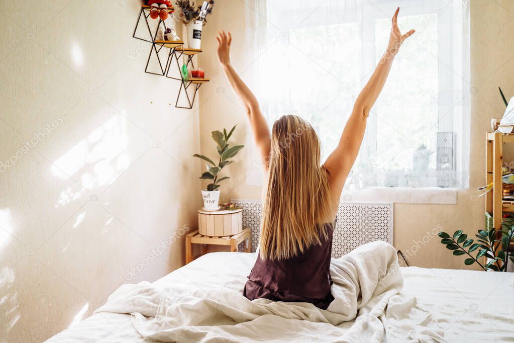 A beautiful blonde stretching sweetly in her bed on an early sunny summer morning. Lazy weekend. The freelancer himself creates the schedule of his day. Back view.