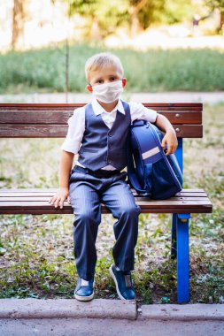 Koruyucu tıp maskeli genç bir öğrenci okulun önündeki bankta oturuyor. Resmi üniforma giymiş, sırt çantası taşıyor. Koronavirüs salgını sırasında yeni bir gerçeklik. Covid 19.