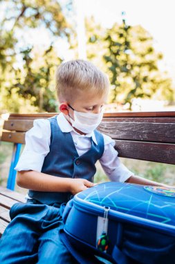 Birinci sınıfın genç öğrencisi okul bahçesinde duruyor. Sırt çantasındaki eşyaları inceliyor. Covid-19 Coronavirus salgını sırasında yeni güvenlik gereksinimleri nedeniyle koruyucu tıbbi maske takıyor..