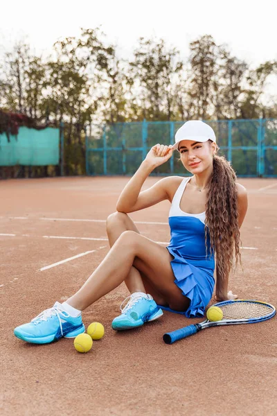 Sports shooting on a clay tennis court. Attractive young girl sexy posing sitting on the court. Dressed in a sports dress and a cap. Tennis racket and balls are scattered nearby.