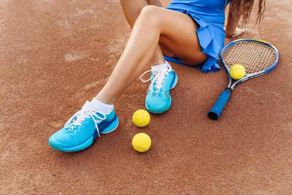 Sports shooting on a clay tennis court. Attractive young girl sexy posing sitting on the court. Dressed in a sports dress and a cap. Tennis racket and balls are scattered nearby.