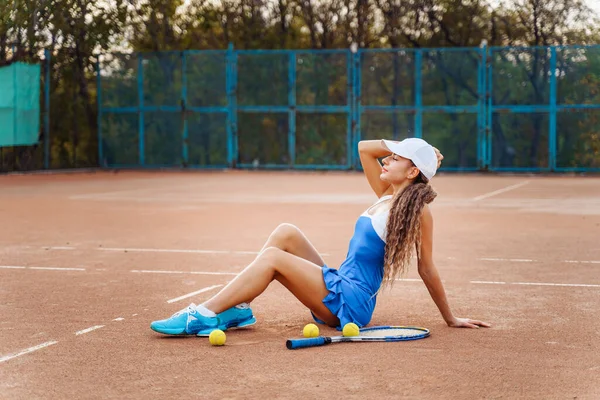 クレイテニスコートでのスポーツシューティング 魅力的な若い女の子のセクシーなポーズは 裁判所に座っている スポーツドレスとキャップに身を包んだ 近くにテニスラケットとボールが散乱しています — ストック写真