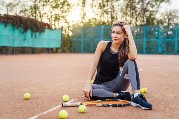 屋外のテニスボールの間でネットテニスコートの近くに座って美しい若い女性の肖像画 スポーツ女性はテニスコートで休んでいる スタイリッシュなスポーツウェアを着たポーズ — ストック写真