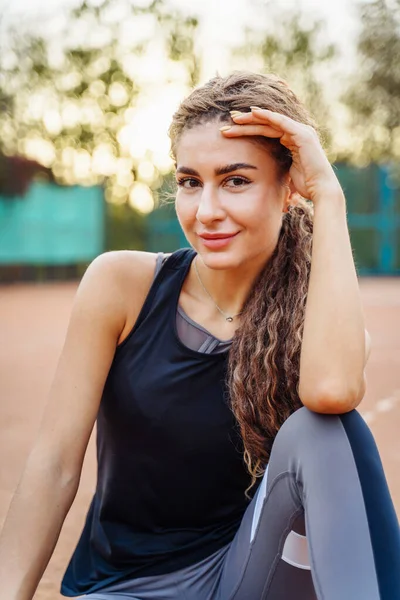 屋外のテニスボールの間でネットテニスコートの近くに座って美しい若い女性の肖像画 スポーツ女性はテニスコートで休んでいる スタイリッシュなスポーツウェアを着たポーズ — ストック写真