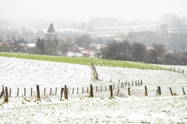 During snowing with a gray sky and snow on the field with a church tower in the middle of the village