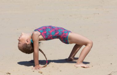 Beş yaşında, kumsalda mayo giymiş, köprü egzersizi yapan beyaz bir kız.