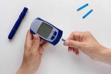 Beyaz arkaplandaki kan şekeri seviyesini hesaplamak için ellerinde glikoz ölçer ve test şeridi tutan kadınların üst görünümü