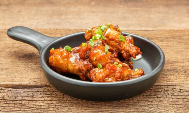 Baharatlı sosla kızartılmış tavuk, beyaz susam ve doğranmış soğan, ahşap bir masada siyah seramik bir tavaya konur..