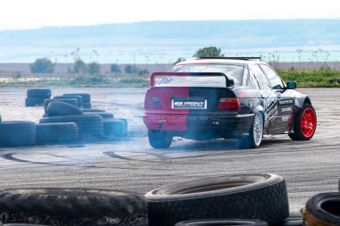 Buhovtsi, Bulgaristan - 12.10.2025 - Siyah ve kırmızı bir araba açık hava pistinde sürükleniyor ve duman bulutları gönderiyor. Lastikler çevreyi çiziyor ve manzara bulutsuz bir gökyüzünün altındaki uzak tepeleri gösteriyor..