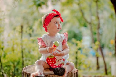 Şirin kırmızı fiyonklu neşeli bir bebek kütüğün üzerinde oturmuş ve bahçede gününün tadını çıkarıyor.
