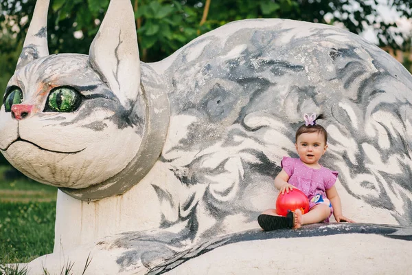 玩世不恭的婴儿 身穿紫色衣服 头戴蝴蝶弓 手持红球 坐在石猫雕像上 — 图库照片