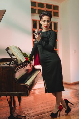 Long shot of stylish and fashionable elegant woman posing for camera at home next to the piano and holding wine glass