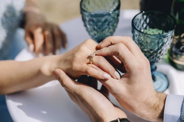Outdoor close up of man proposing to his girlfriend. Guy puts engagement ring and leaning elbows on table with glasses