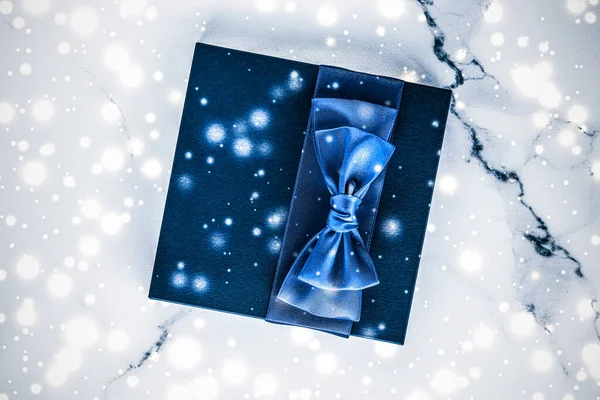 Winter holiday gift box with blue silk bow, snow glitter on marble background as Christmas and New Years presents for luxury beauty brand, flatlay design