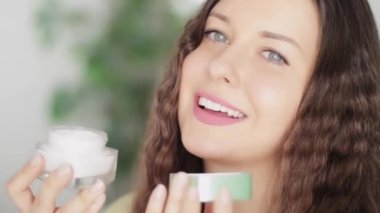 Güzel gülümseyen genç bir kadın açık yüz kremi kavanozunu tutuyor, doğal güzellik olarak organik kozmetik ürünü ve günlük sağlıklı cilt bakımı rutini, lüks kozmetik ürünleri, insanlar ve sağlık reklamı.