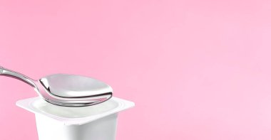 Yoğurt kabı ve pembe arka planda gümüş kaşık yoğurt kremalı beyaz plastik kap sağlıklı beslenme ve beslenme dengesi için taze süt ürünü.