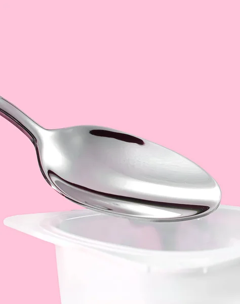 Copa de yogur y cuchara de plata sobre fondo rosa, recipiente de plástico blanco con crema de yogur, productos lácteos frescos para una dieta saludable y equilibrio nutricional —  Fotos de Stock