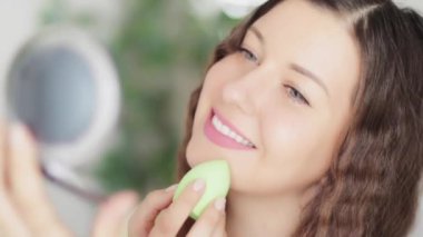 Güzel bir kadın doğal organik makyaj uyguluyor. Güzellik blenderi kullanıyor, gülümsüyor, eko makyaj malzemesi kullanıyor, beyaz modelin ya da vlogger 'ın yüz portresi kozmetik ürün, cilt bakımı ve insan konsepti kullanıyor.