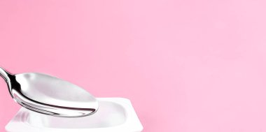 Yoğurt kabı ve pembe arka planda gümüş kaşık yoğurt kremalı beyaz plastik kap sağlıklı beslenme ve beslenme dengesi için taze süt ürünü.