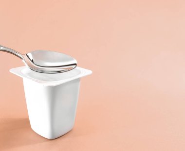 Yoğurt kabı ve bej arka planda gümüş kaşık, yoğurt kremalı beyaz plastik kap, sağlıklı beslenme ve beslenme dengesi için taze süt ürünü