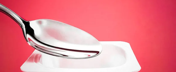 Copa de yogur y cuchara de plata sobre fondo rojo, recipiente de plástico blanco con crema de yogur, productos lácteos frescos para una dieta saludable y equilibrio nutricional — Foto de Stock